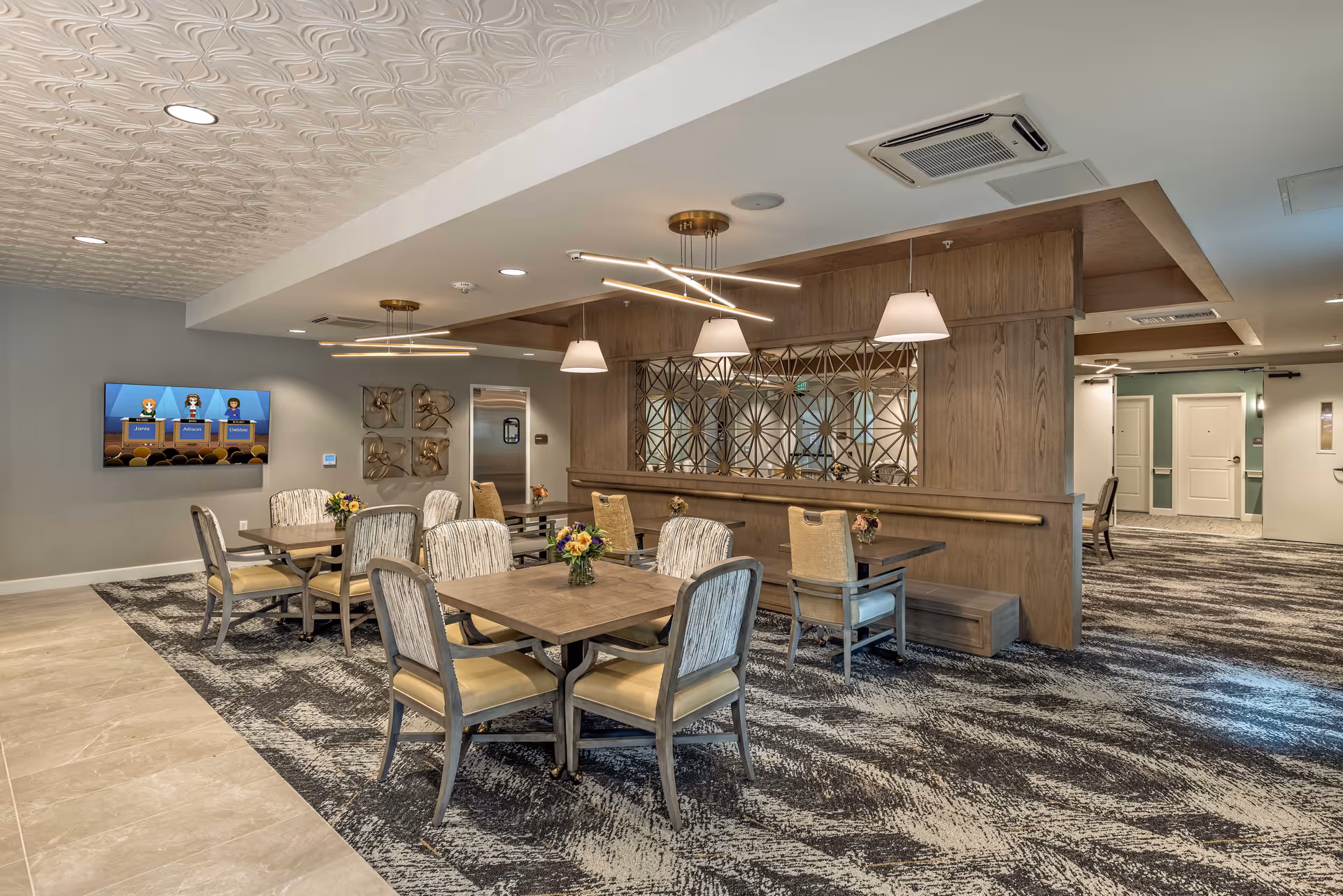 A modern dining area in a senior living facility featuring several square wooden tables with cushioned chairs arranged around them. The room has a patterned carpet and tiled floor, decorative wall art, a large mirror with a geometric design, and contemporary ceiling light fixtures. A flat-screen TV is mounted on the wall displaying a game show with animated characters.