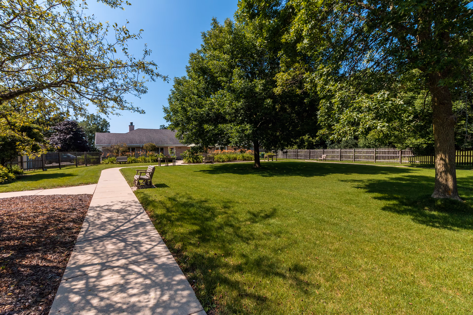 Sunny grassy courtyard with a concrete walkway, benches, mature trees, and a building in the background.