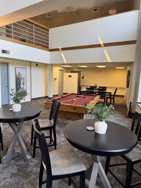 Interior common area of St. Catherine Retirement Community featuring round tables with chairs and potted plants, a pool table with cues, and a dining table with chairs in the background. The space has a high ceiling with a second-floor balcony and neutral-colored walls and carpet.