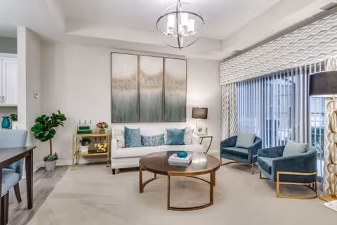 A modern living room with a white sofa adorned with blue and white pillows, two blue armchairs with gold frames, a round wooden coffee table with books and a decorative item, a gold bar cart with plants and decor, a large abstract triptych painting on the wall, a floor lamp with a black shade, and large windows with vertical blinds and patterned valance.