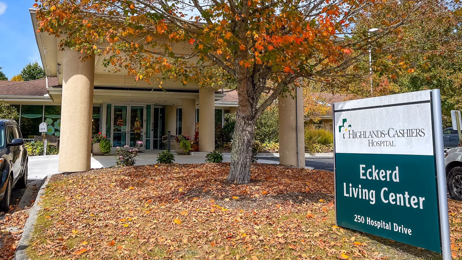 Exterior view of the entrance to Eckerd Living Center, part of Highlands-Cashiers Hospital, with a large sign displaying the facility name and address. The entrance is framed by large columns and surrounded by autumn foliage and parked cars.