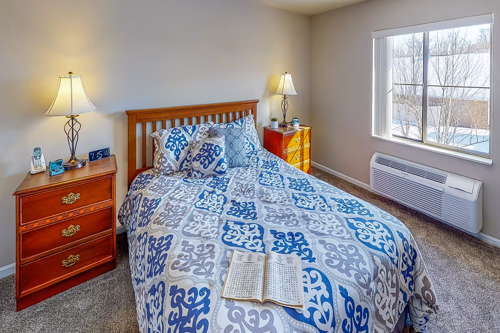 A bedroom with a wooden bed frame and a bed covered in a blue, gray, and white patterned comforter. There are multiple pillows on the bed, and an open book is lying on the comforter. On each side of the bed are wooden nightstands with lamps, a clock, a phone, and a small plant. A window with vertical blinds shows a snowy outdoor scene, and a wall-mounted air conditioning unit is below the window.