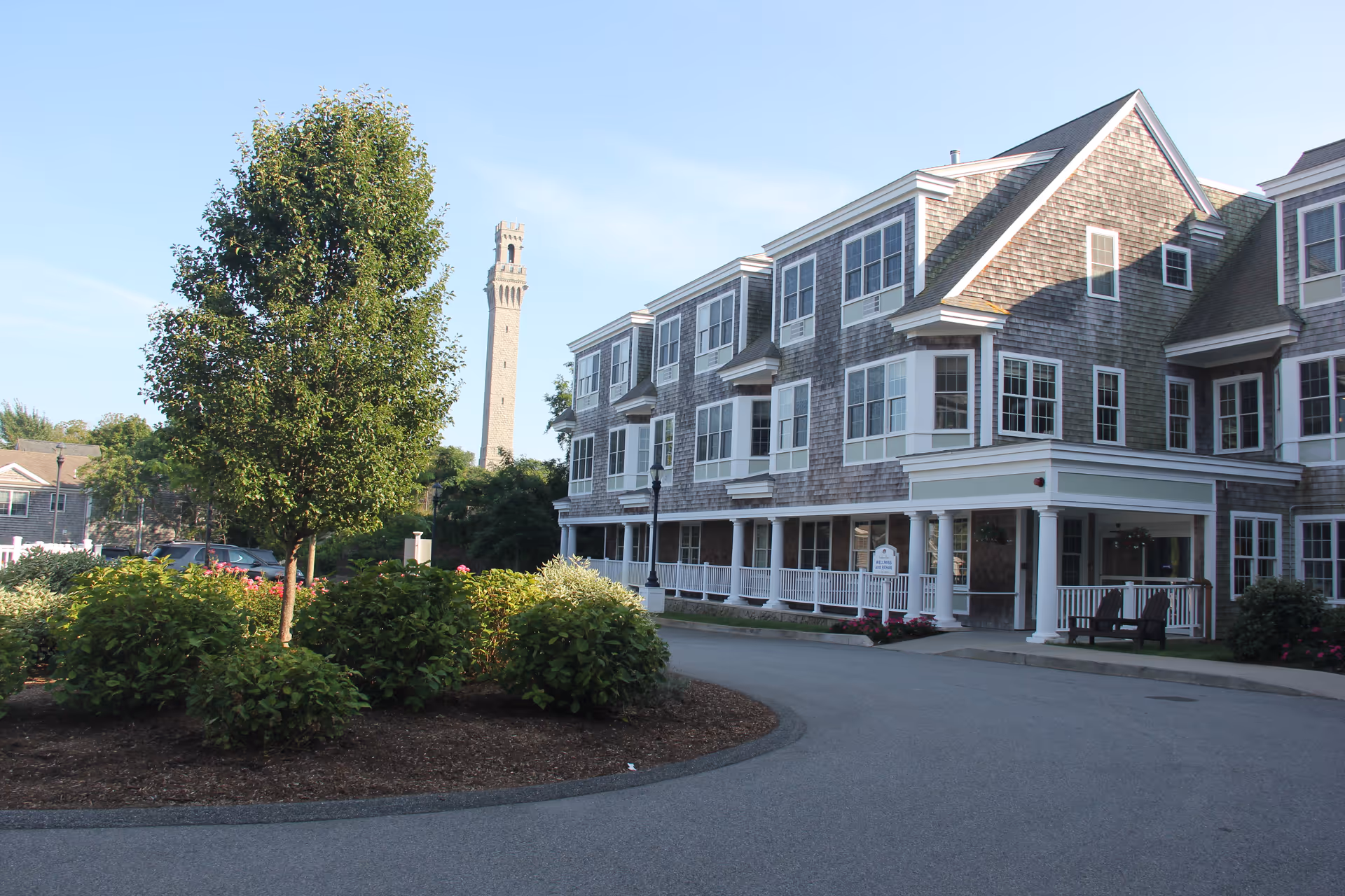 Exterior view of a multi-story senior living facility building with gray shingles and white trim. There is a curved driveway in front with landscaped bushes and a tree in the center. A tall tower structure is visible in the background under a clear blue sky.