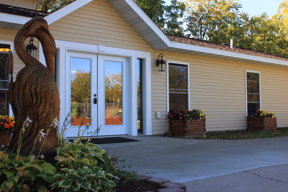 Exterior view of a single-story building with beige siding, white-framed double glass doors, two windows, and flower planters beneath the windows. A wooden sculpture of a bird is visible in the foreground surrounded by greenery and flowers.