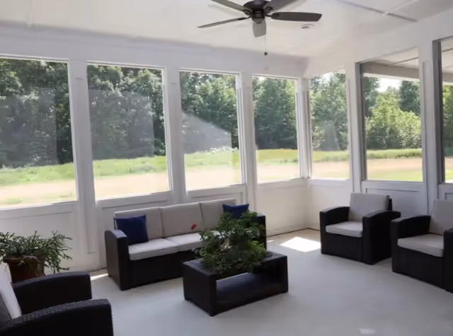 A bright enclosed porch or sunroom with large windows showing a green outdoor landscape. The room contains dark wicker furniture with light cushions, including two armchairs, a loveseat, and a rectangular coffee table with a potted plant. A ceiling fan is mounted on the white ceiling.