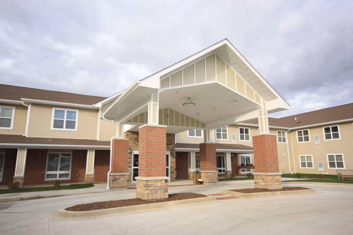 Entrance of a senior living facility with a covered drop-off area supported by brick and stone pillars. The building has beige siding with white trim and multiple windows. The sky is cloudy.