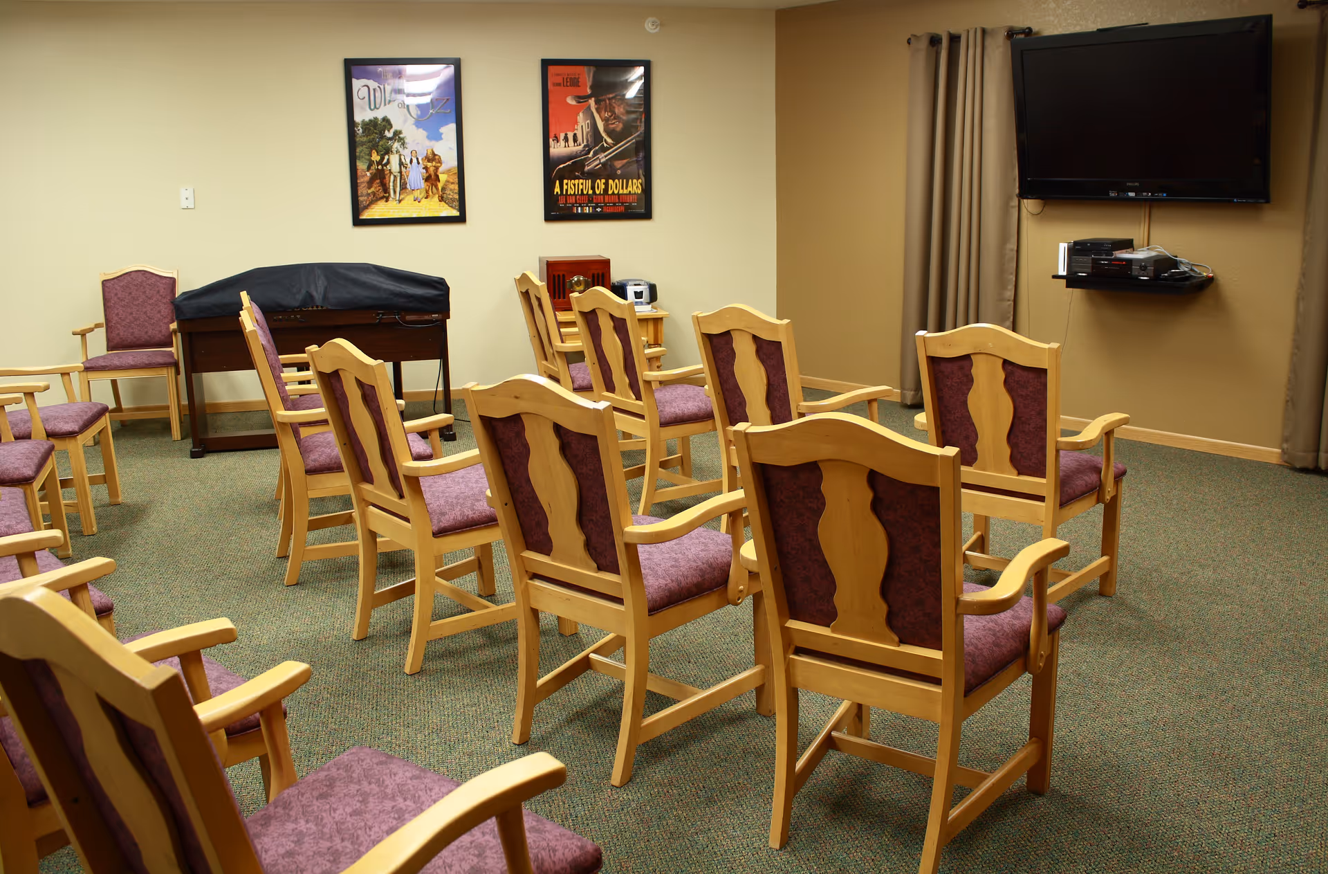 A small meeting or activity room with wooden chairs that have purple cushions arranged in rows facing a wall-mounted flat screen TV. The room has beige walls, carpeted floor, two framed movie posters on the wall, and a covered piano or organ in the corner.