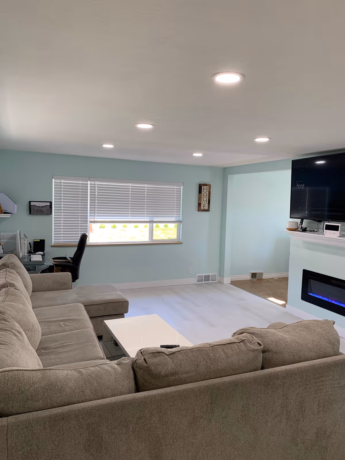 A bright living room with light blue walls and white flooring. There is a large gray sectional sofa with a white coffee table in front of it. On the right wall, there is a mounted flat-screen TV above a modern electric fireplace with blue flames. A window with white blinds is on the far wall, and a black office chair and desk with a computer are positioned near the window. A small decorative sign that reads 'HOME' is hung on the wall near the window.