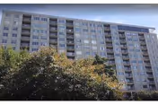 Exterior view of a multi-story residential building with numerous windows and balconies, partially obscured by trees in the foreground under a clear sky.