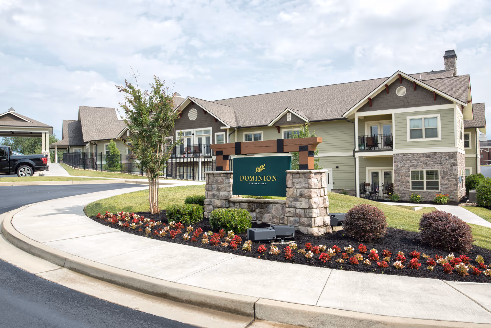 Exterior view of Dominion Senior Living facility showing a landscaped entrance with a stone sign, flower beds, and a two-story building with balconies and a covered parking area.