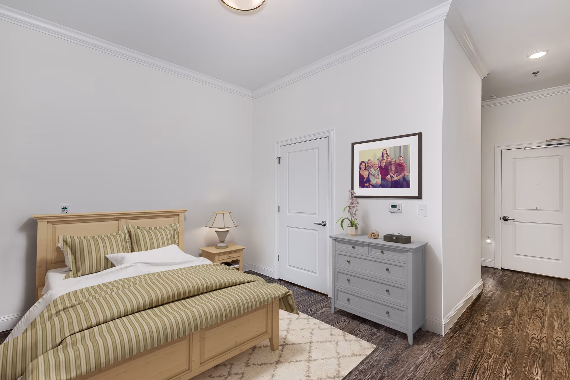 A bedroom with a wooden bed frame and striped bedding in shades of green and beige. There is a matching wooden nightstand with a lamp on it next to the bed. Across from the bed is a gray dresser with decorative items and a framed family photo hanging above it. The room has white walls, dark wood flooring, and a light-colored area rug. A hallway with two white doors is visible to the right.