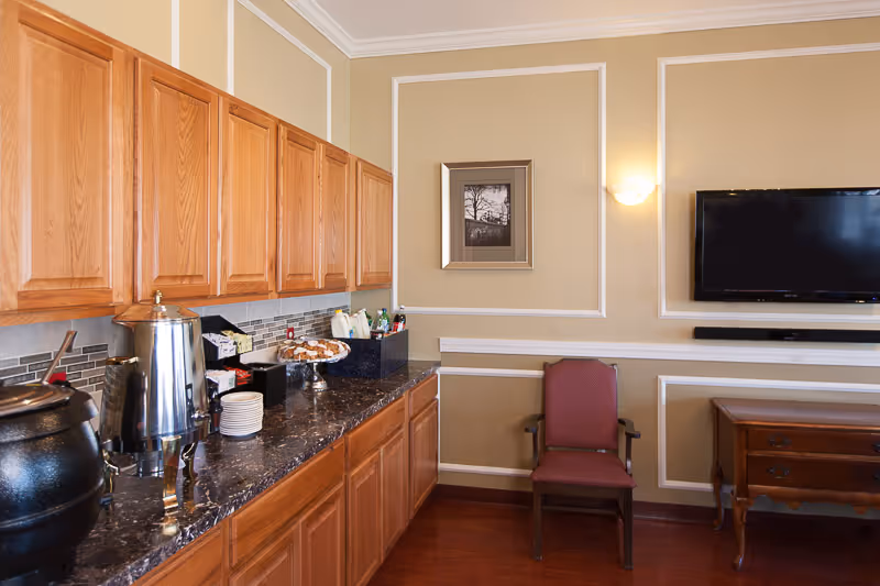 Interior view of a room with wooden cabinets and a dark marble countertop on the left side, holding a large metal beverage dispenser, a soup pot, plates, and a tray of pastries. On the right side, there is a red upholstered chair, a wooden side table, a wall-mounted flat screen TV, and a framed black and white photograph on the beige wall with white trim.