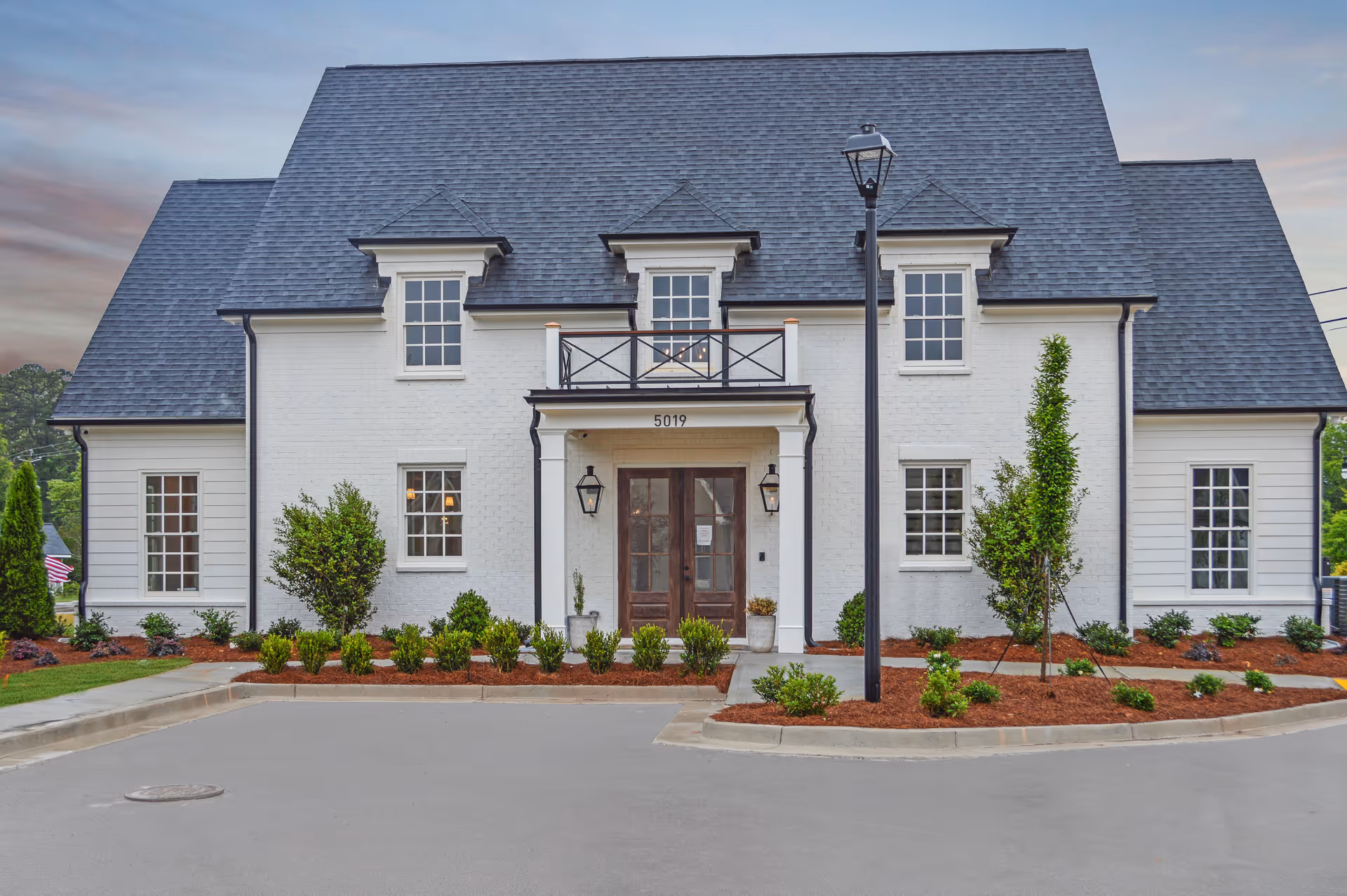 Front exterior view of a white two-story building with a dark gray roof, multiple windows, a central entrance with double wooden doors, and a small balcony above the entrance. The building is surrounded by landscaped bushes and small trees, with a paved driveway in front.
