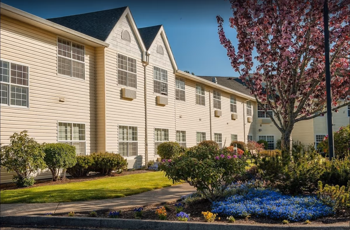 Front exterior of a two-story senior living building with landscaped gardens, flowering shrubs, and a sidewalk.