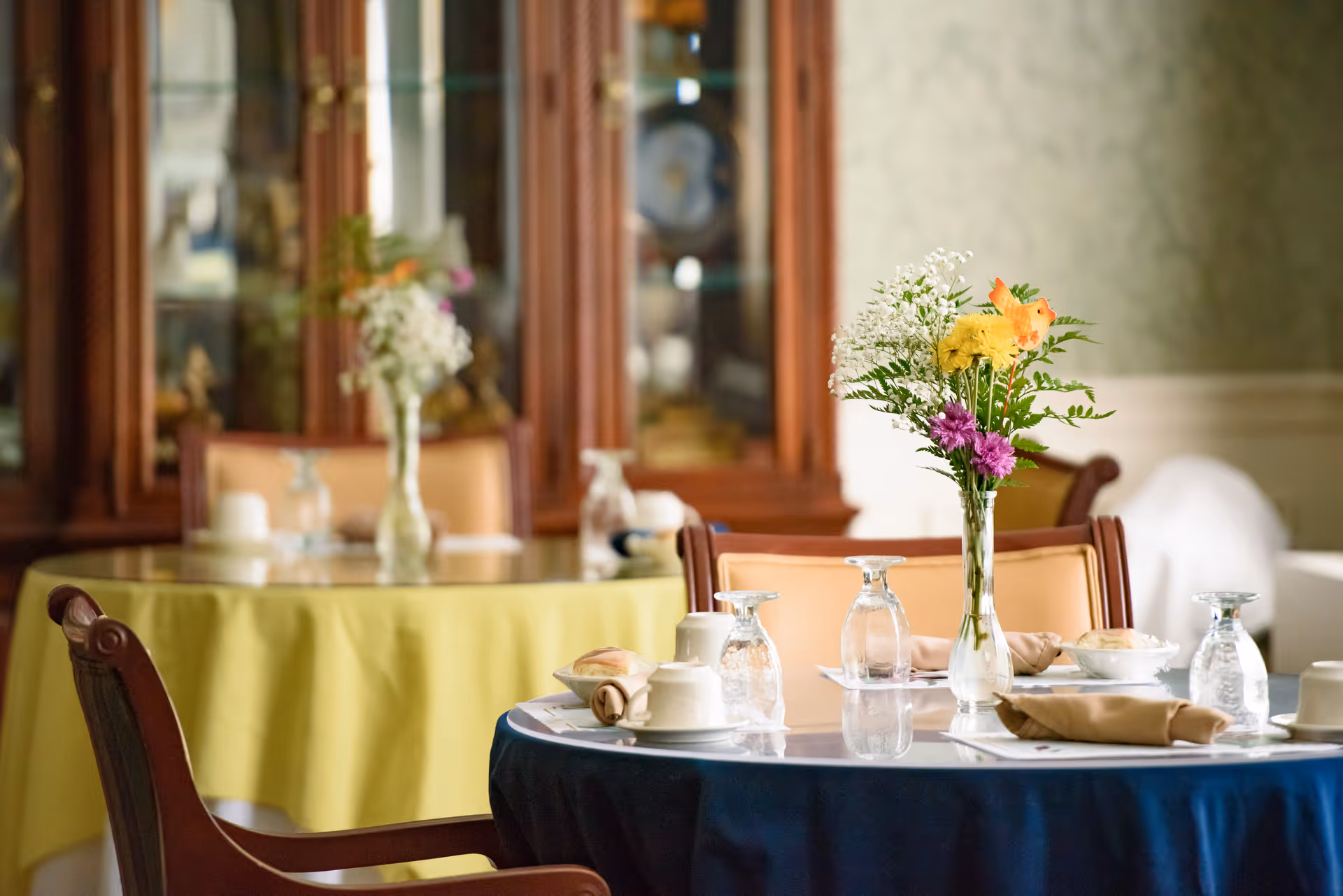 A dining room with tables covered in blue and yellow tablecloths, set with glasses, cups, napkins, and a vase of colorful flowers on each table. In the background, there is a wooden cabinet with glass doors displaying decorative items.