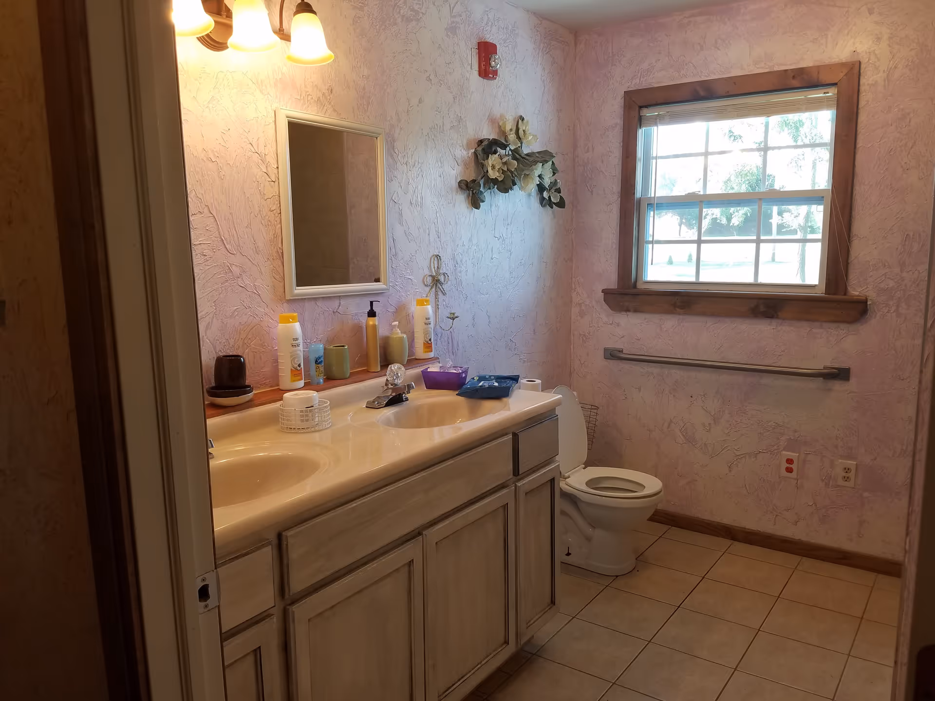 A bathroom with a double sink vanity, a mirror above the sinks, and various toiletries on the countertop. There is a toilet next to the vanity, a window with wooden trim letting in natural light, and a grab bar on the wall beside the toilet. The walls have a textured, light pink finish and the floor is tiled.