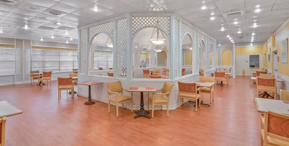 A spacious dining room in a senior living community with multiple small tables and chairs arranged neatly. The room features wooden flooring, white lattice partitions with arches, and large windows with blinds allowing natural light to enter. The ceiling has recessed lighting and a hanging light fixture above one of the tables.