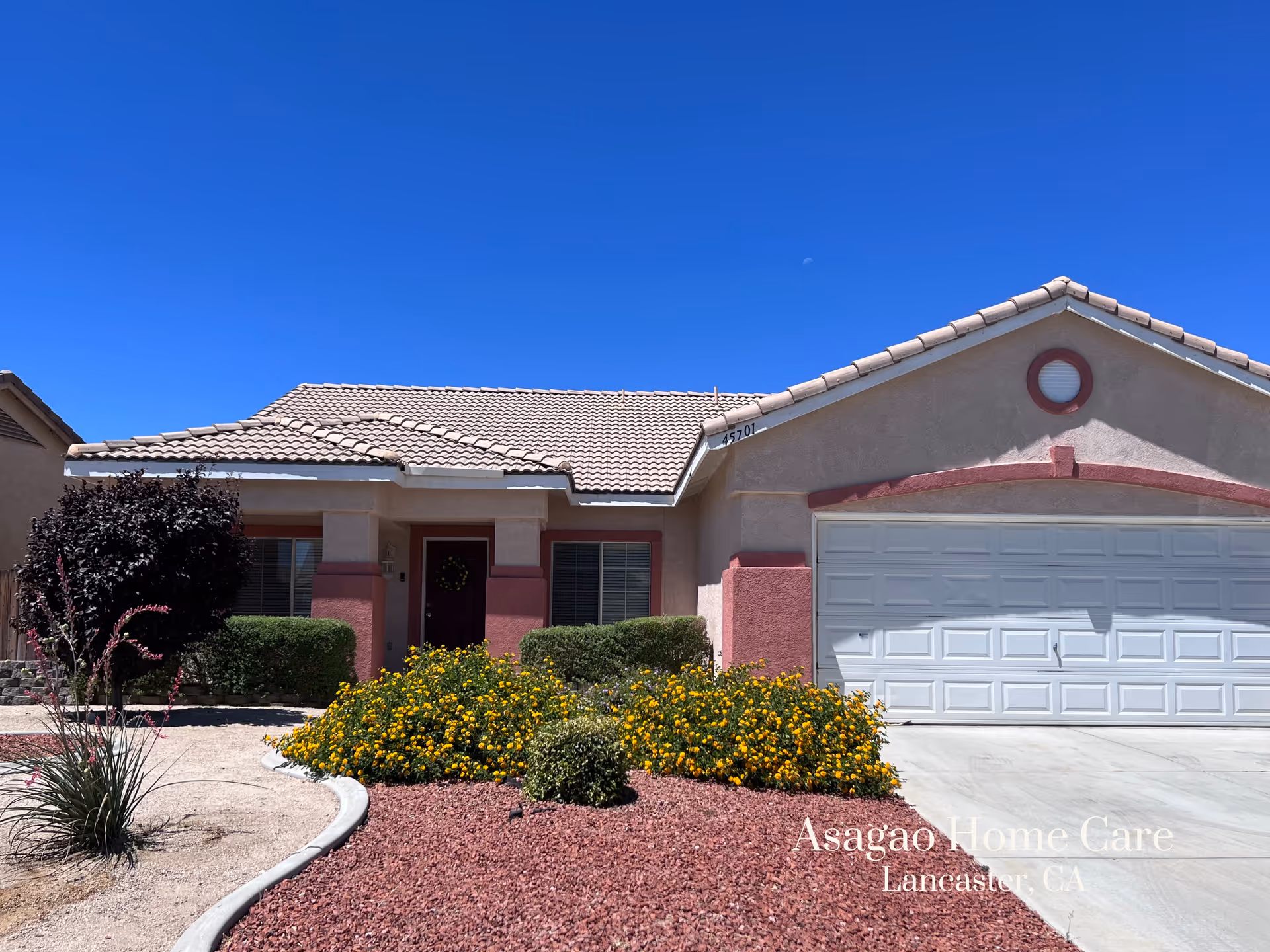 Single-story stucco house with a two-car garage, tiled roof, and flowering shrubs in the front yard under a clear blue sky.