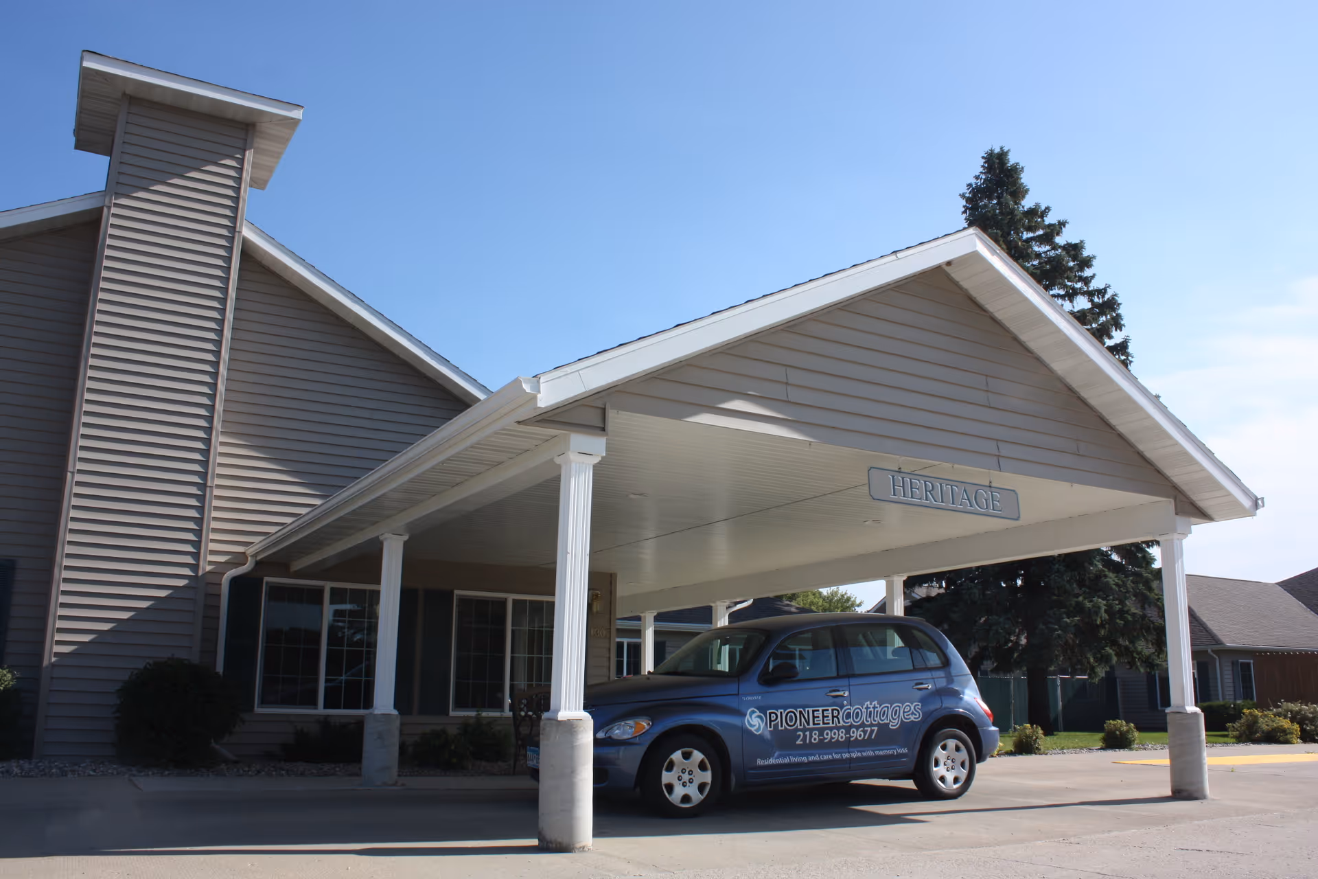 Exterior view of a senior living facility named Heritage with a covered entrance supported by white columns. A blue car with the Pioneer Cottages logo and contact number is parked under the entrance. The building has beige siding and several windows, with a large tree visible in the background under a clear blue sky.