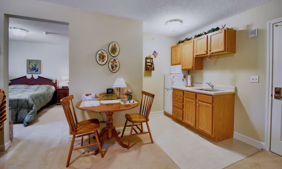 A small apartment interior featuring a kitchenette with wooden cabinets, a white refrigerator, microwave, and sink. Adjacent to the kitchenette is a round wooden table with two wooden chairs. On the wall above the table are three decorative plates and a small lamp on the table. In the background, a bedroom with a bed covered in a green comforter and a nightstand with a lamp is visible.
