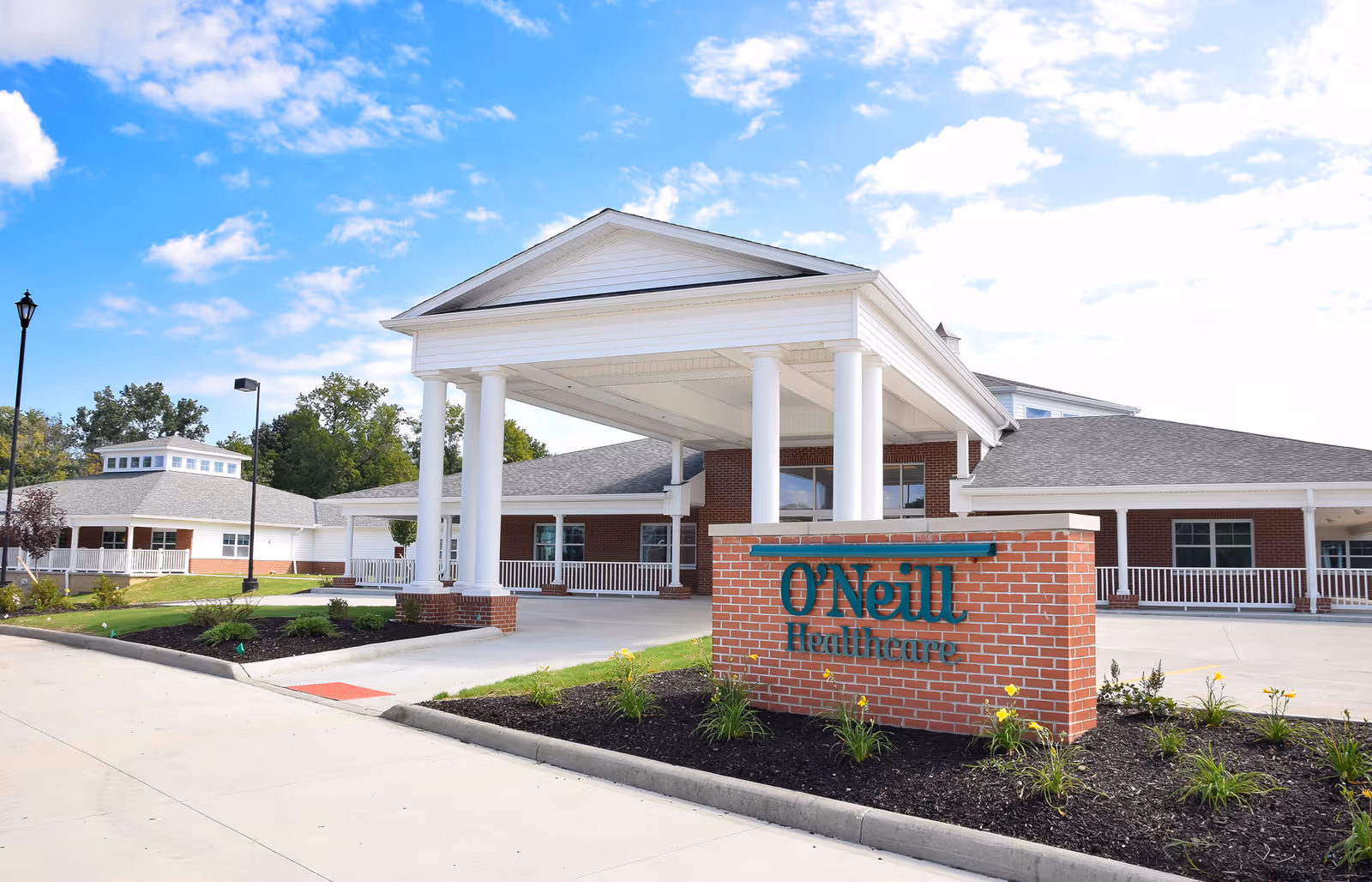 Exterior view of O'Neill Healthcare Fairview Park building with a covered entrance supported by white columns, a brick sign with the facility name, and a clear blue sky with some clouds.
