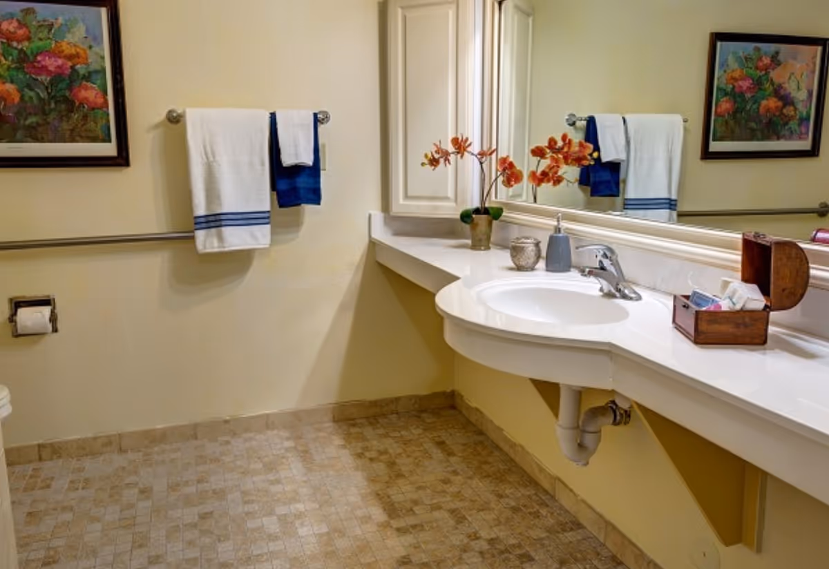 A clean bathroom with a white countertop and sink, a large mirror above the sink, a small vase with orange flowers, a soap dispenser, and a wooden box with toiletries. Two towels, one white with blue stripes and one solid blue, hang on a towel rack on the wall. There is a toilet paper holder with a roll of toilet paper and a framed floral painting on the wall. The floor is tiled in a beige pattern.