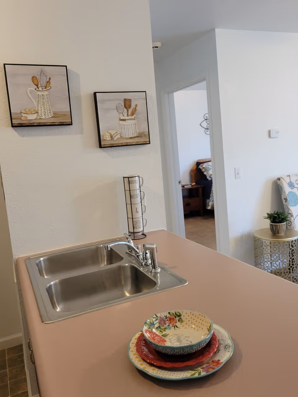 Kitchen countertop with a double stainless steel sink, stacked colorful bowls, and decorative wall art beside an open doorway into another room.