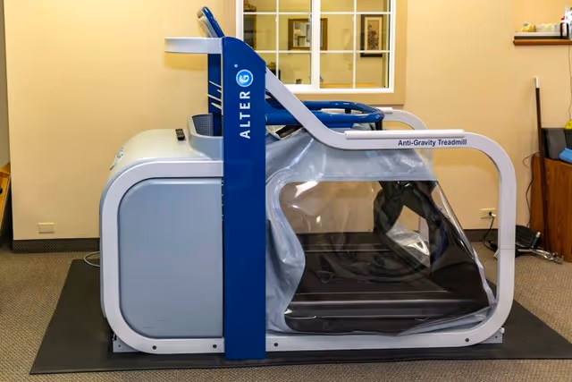An AlterG Anti-Gravity Treadmill in a rehabilitation room with beige walls and carpeted floor, featuring a window and some furniture in the background.
