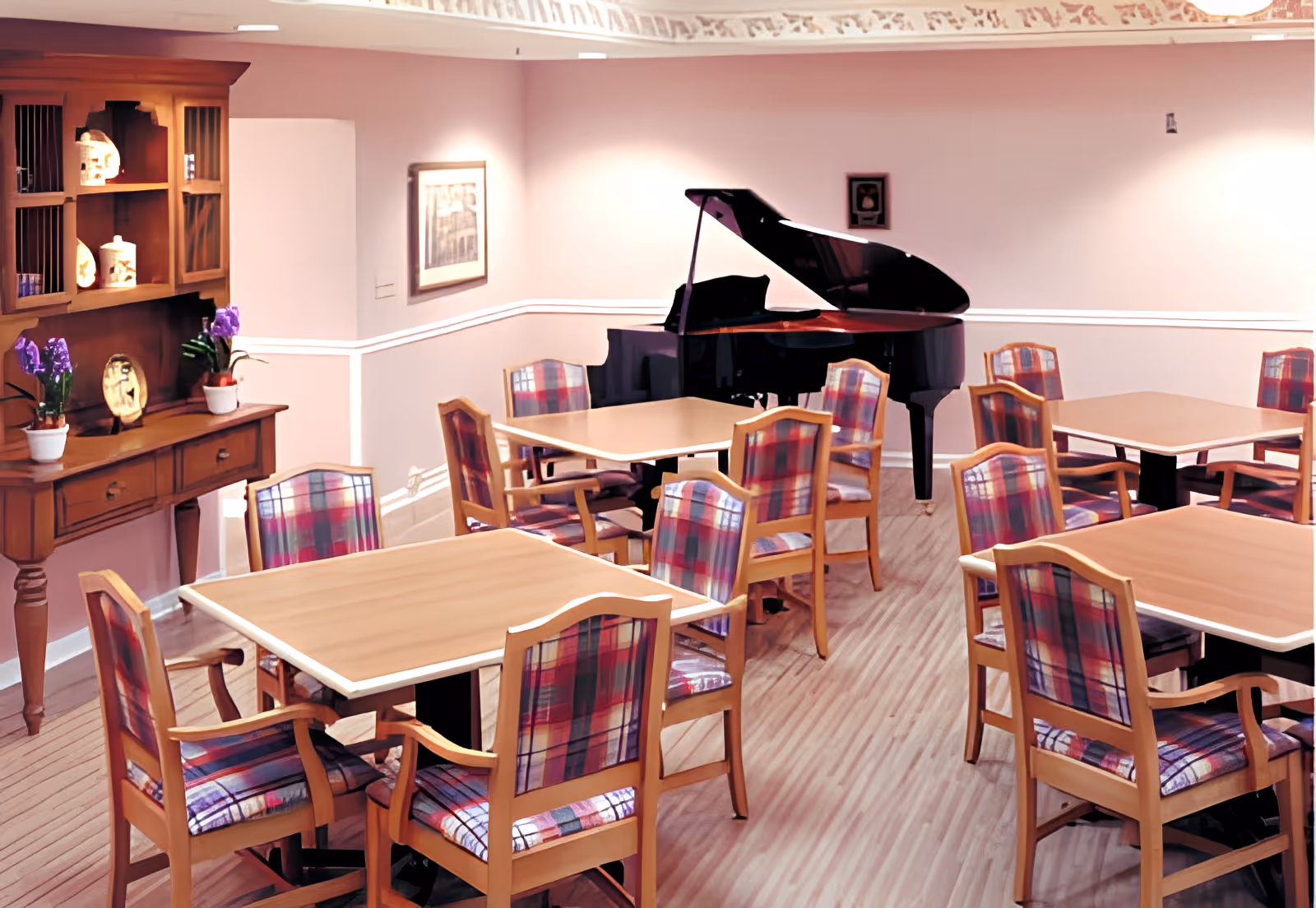 A dining room with several wooden tables and chairs featuring plaid upholstery. A black grand piano is positioned against the far wall. There is a wooden hutch with decorative items and potted plants on the left side. The walls are painted light pink with a white chair rail, and the floor has a light wood finish.
