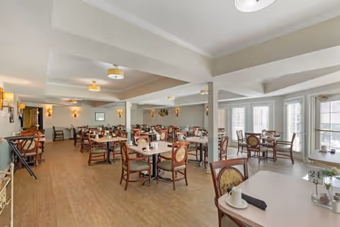 A spacious dining room in an assisted living facility with multiple tables and chairs arranged neatly. The room features large windows allowing natural light to fill the space, light-colored walls, and ceiling lights. Each table is set with cups, napkins, and small decorative items.