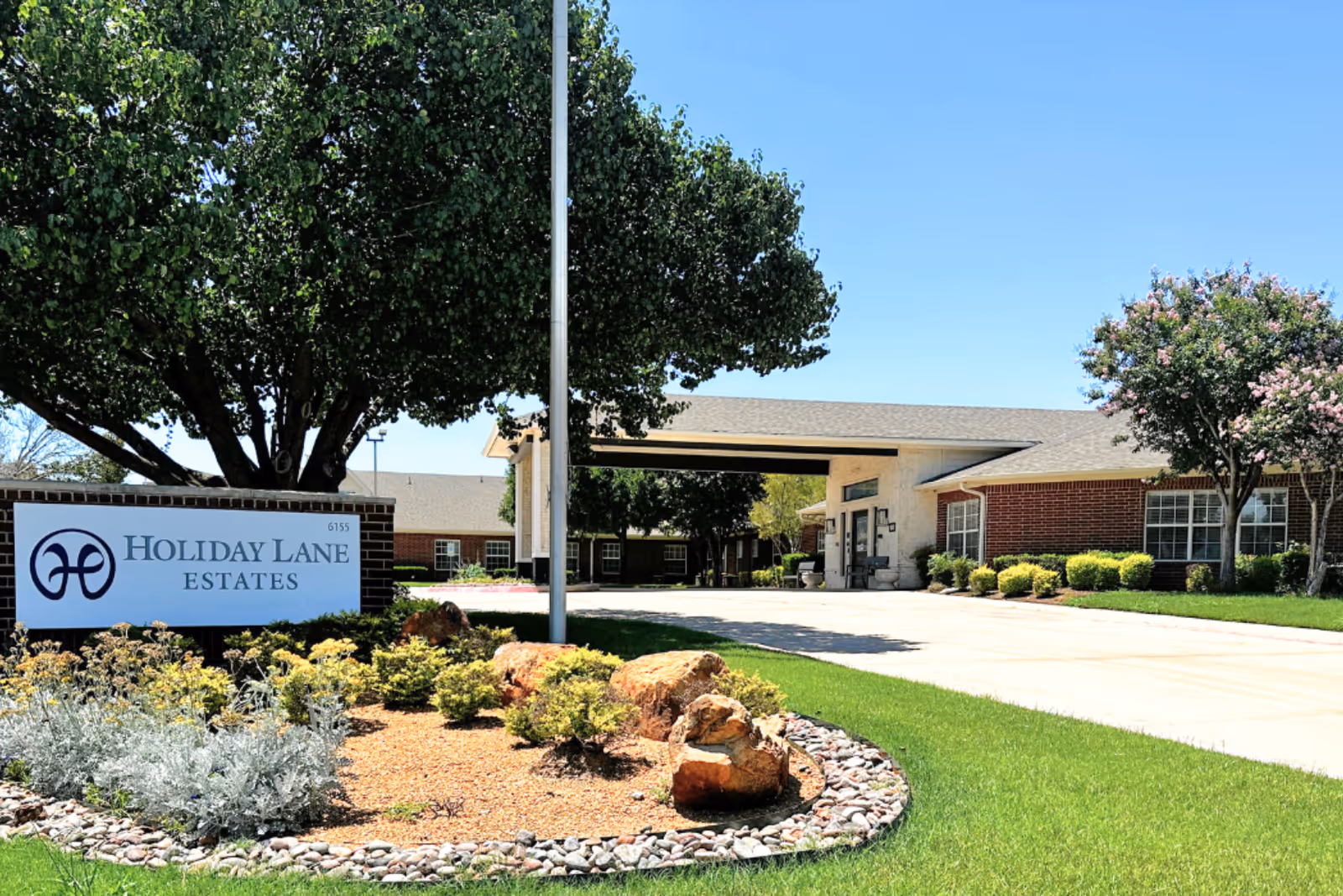 Exterior view of Holiday Lane Estates senior living facility with a driveway, landscaped garden with rocks and shrubs, and a large tree. The building has a covered entrance and brick walls under a clear blue sky.