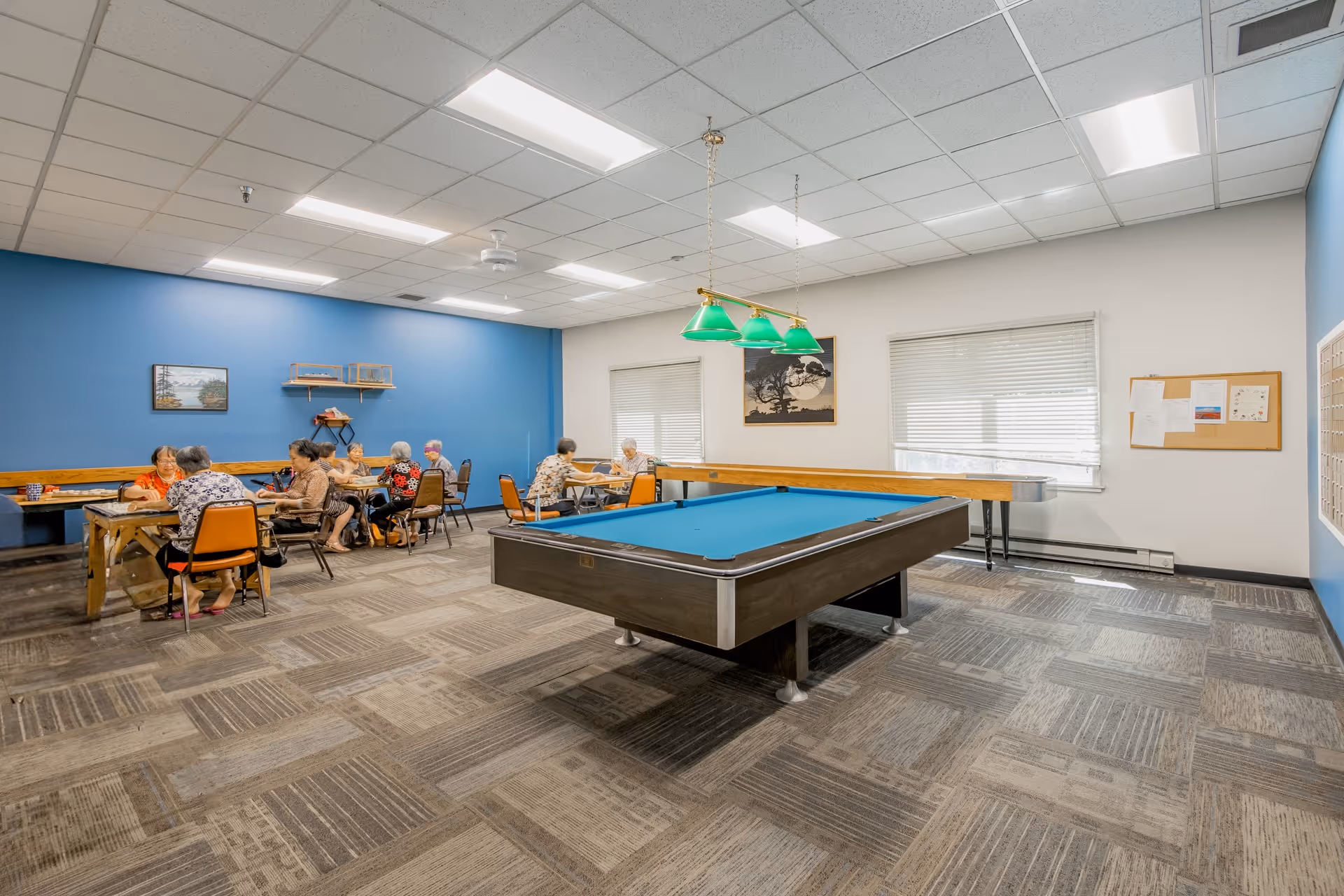 A community activity room with a pool table in the center and several seniors seated at tables playing games.
