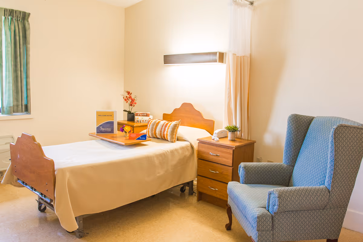 A simple nursing home bedroom with a single bed covered in a beige blanket, a wooden headboard, a bedside table with a small plant and a telephone, a blue upholstered armchair, and a window with green curtains. There is a tray on the bed with a welcome book and some flowers.