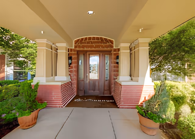 Covered entrance to a building with a wooden double door framed by brick and beige columns, flanked by potted plants and greenery on both sides.