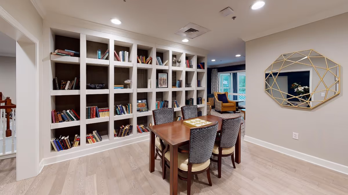 Bright communal lounge with built-in bookshelves, a wooden table and four chairs, and a seating area beyond.