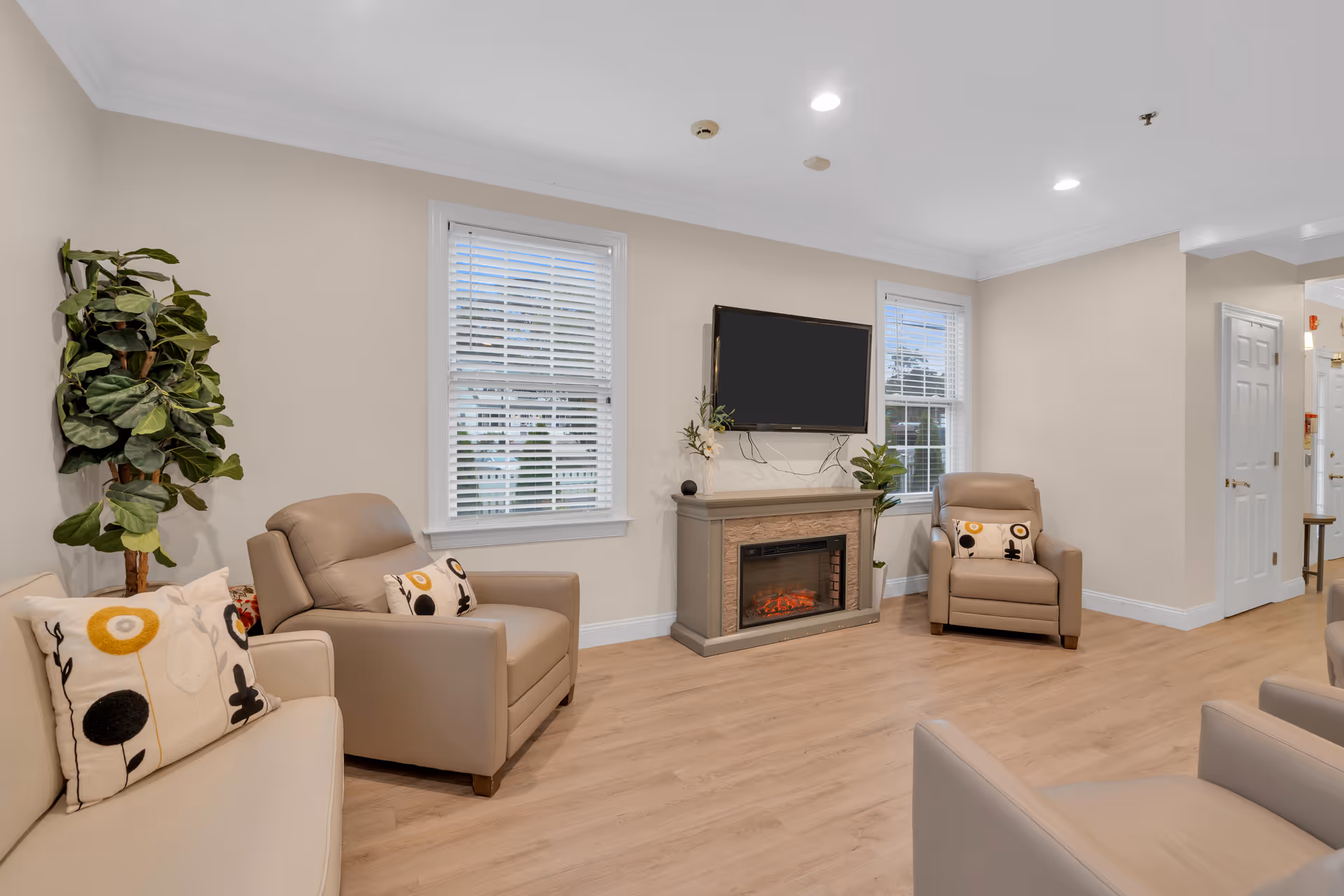 A cozy living room area with beige walls and light wood flooring. The room features a beige sofa and two beige armchairs, each with decorative pillows. There is a flat-screen TV mounted above an electric fireplace, flanked by two windows with white blinds. A tall green plant is placed in the corner near the sofa.