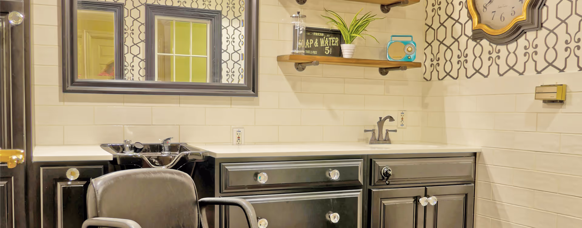 Interior view of a salon area with a black salon chair in front of a black sink and black cabinets with white countertops. A large mirror hangs on the wall above the sink, and a wooden shelf holds a small plant, a vintage-style radio, and a decorative sign. The walls have white tiles on the lower half and patterned wallpaper on the upper half. A clock is mounted on the wall to the right.