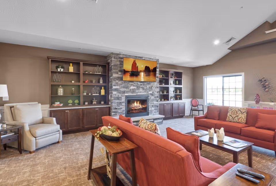 A bright common room with red sofas facing a stone fireplace and TV, built-in wood shelving, armchairs, and a large window.