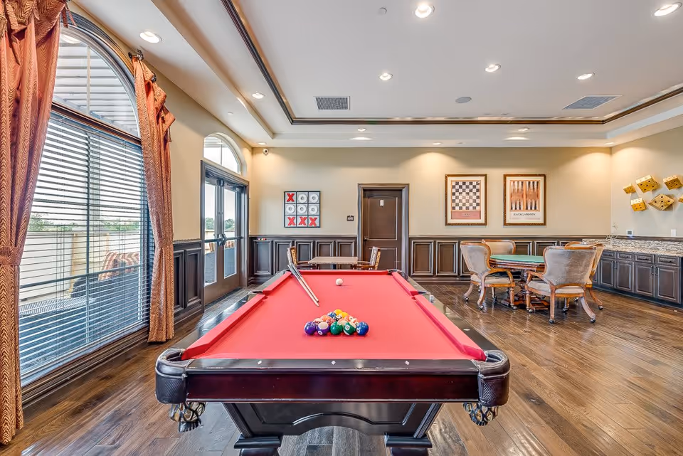 A bright and spacious game room with a red felt pool table in the center, a set of pool balls arranged for a game, and a cue stick resting on the table. The room features large arched windows with brown curtains, a door leading outside, wooden flooring, and several framed game-themed artworks on the walls. There is also a card table with four upholstered chairs and a countertop with cabinets along one wall.