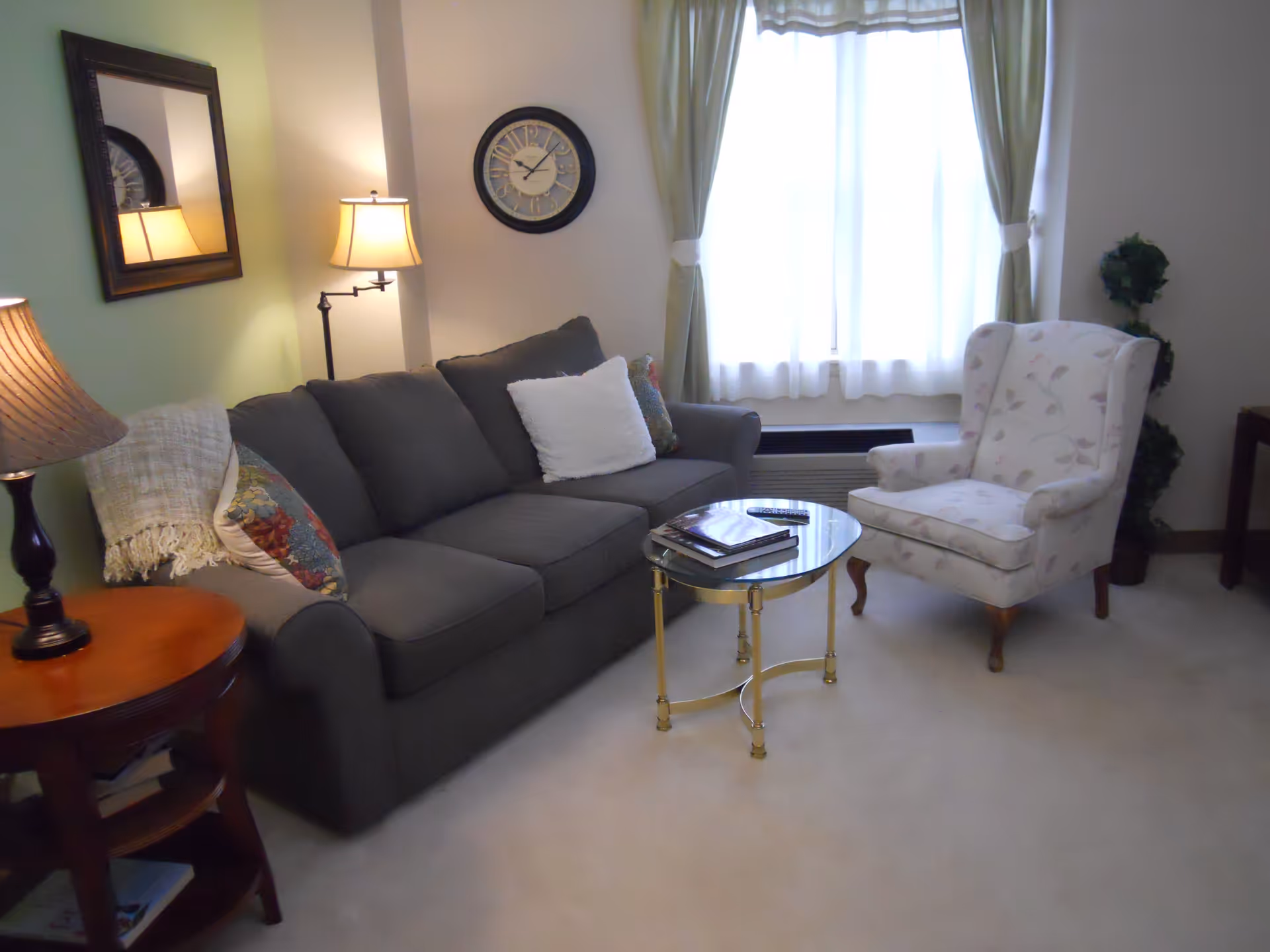 A cozy living room with a dark gray sofa adorned with colorful and white pillows, a floral patterned armchair, a round glass coffee table with books and a remote control, a wooden side table with a lamp, a wall clock, a mirror, and a window with green curtains letting in natural light.