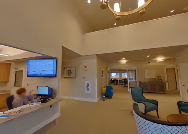 Interior view of a senior living facility reception area with a front desk where a person is seated working on a computer. The space has beige walls and carpet, a chandelier hanging from the ceiling, and several chairs arranged in a seating area. There is a hallway leading to other rooms in the background.