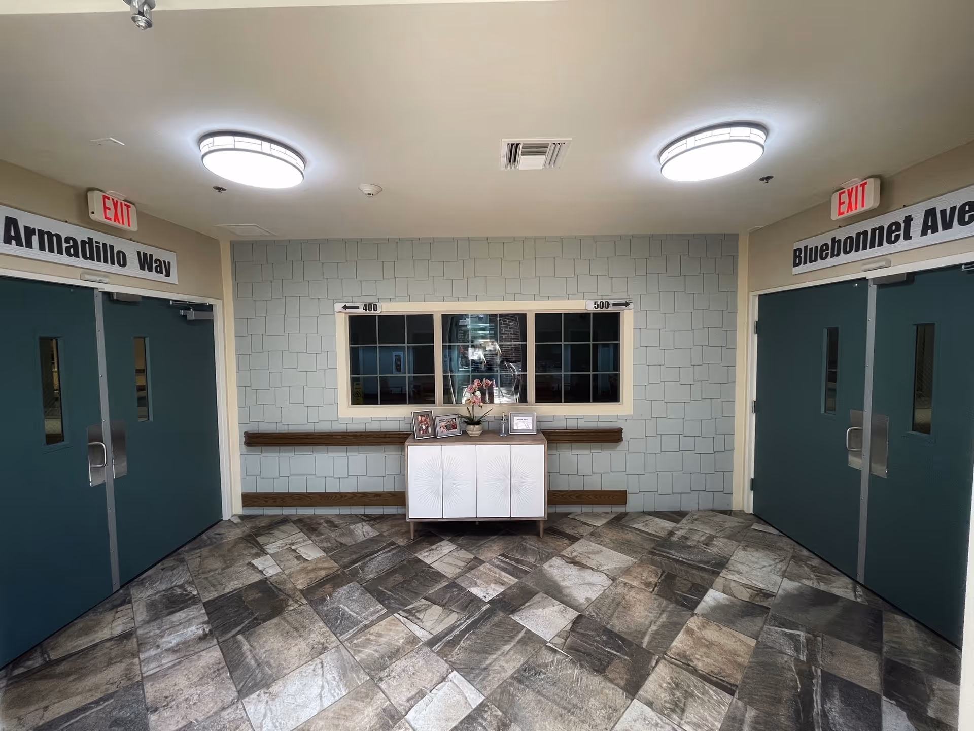 Interior hallway area with two sets of double doors labeled Armadillo Way on the left and Bluebonnet Ave on the right. Between the doors is a light blue shingled wall with a window and a small white cabinet below it, decorated with framed photos and a small plant. The floor is tiled with a mix of gray and beige tones, and two round ceiling lights illuminate the space.