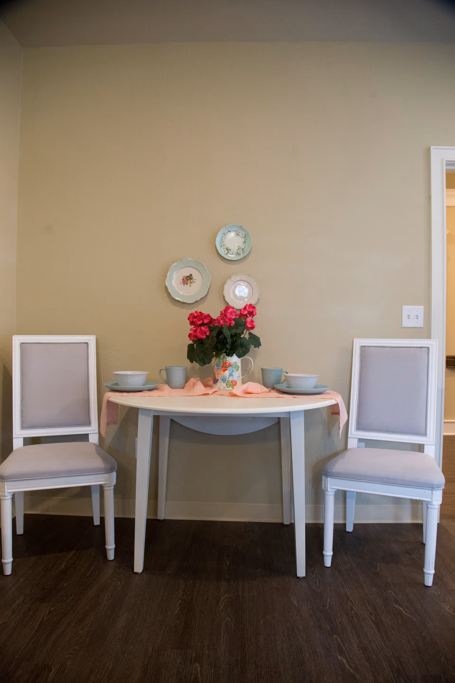 A small dining area with a white table set against a beige wall. The table has a pink table runner, a floral pitcher with bright pink flowers, two place settings with bowls and mugs, and two white chairs with light gray cushions on either side. Three decorative plates are hung on the wall above the table.