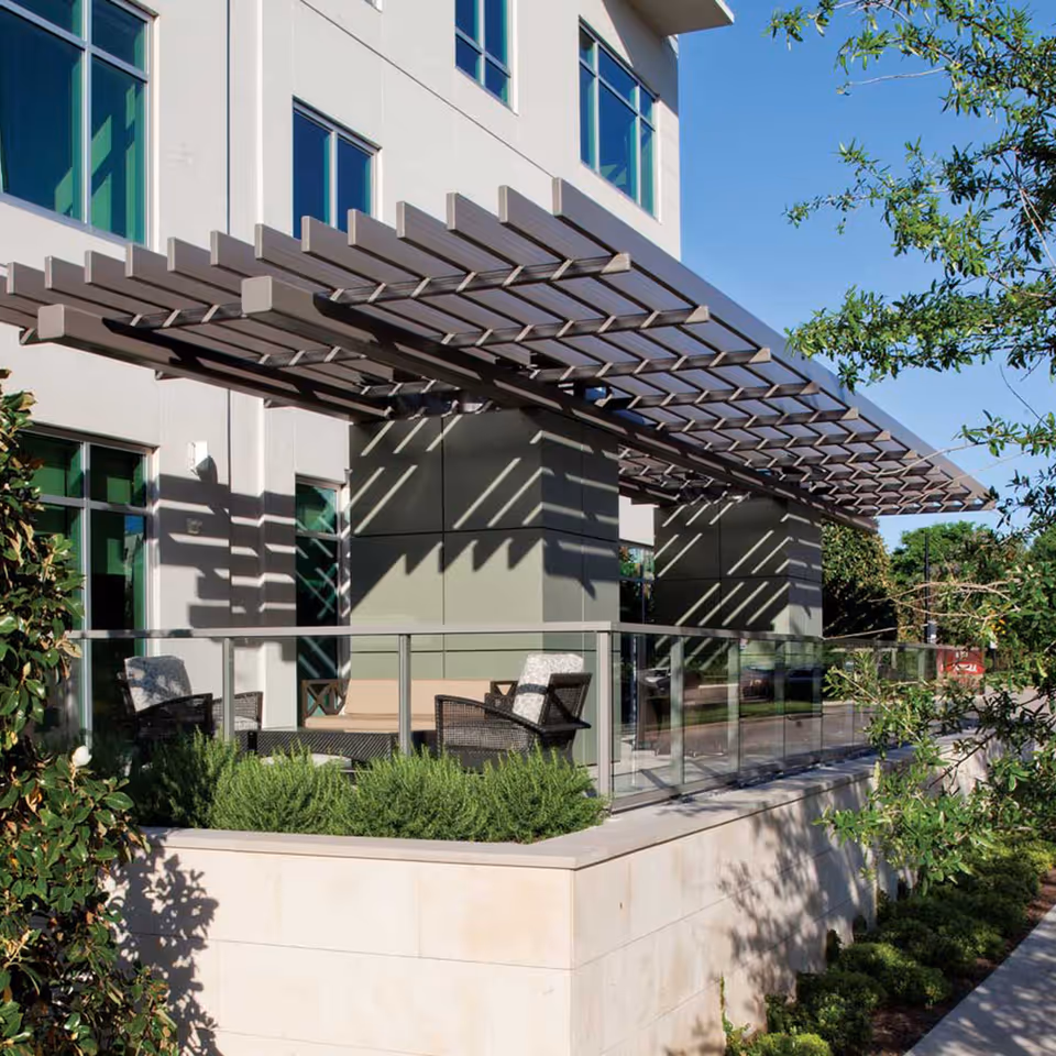 Outdoor seating terrace with a pergola-style awning, glass railing, and chairs along the front of a modern building.