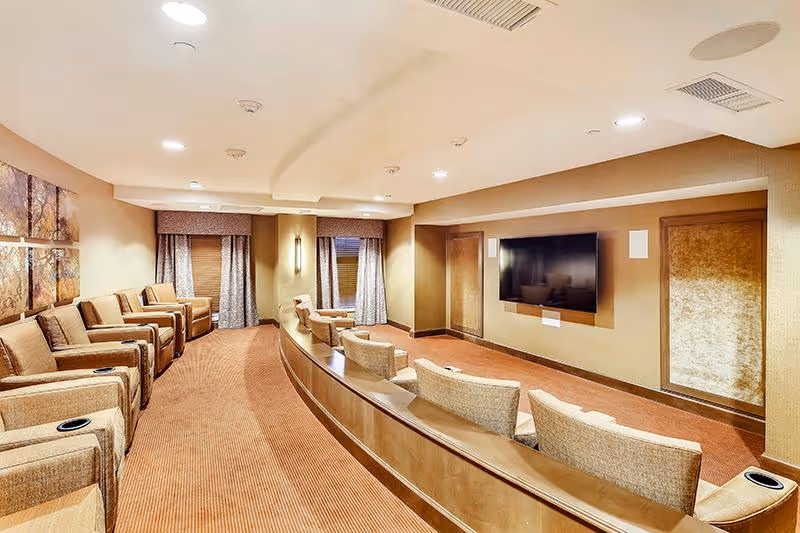 Carpeted community media room with tiered upholstered recliner seating facing a wall-mounted flat-screen television.