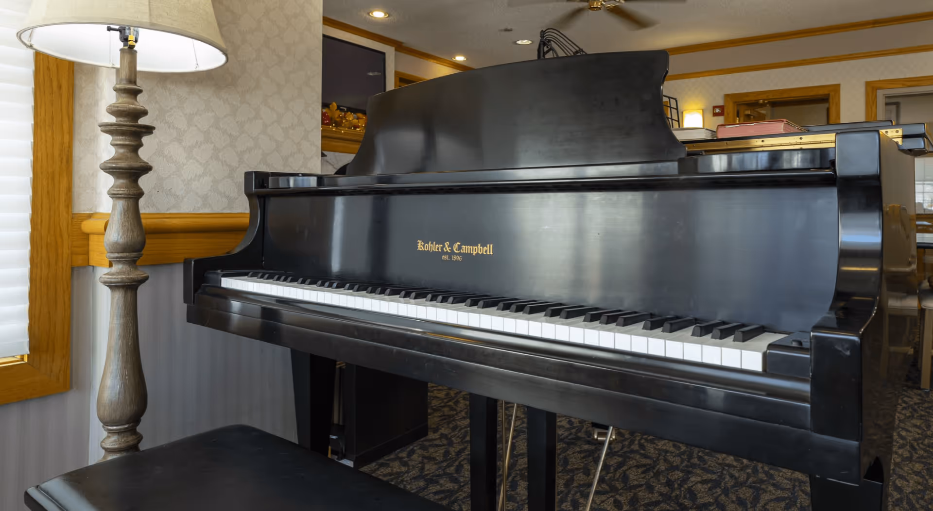 A black Kohler & Campbell grand piano in a room with patterned carpet and wallpaper. A wooden lamp with a white lampshade is positioned to the left of the piano, and a piano bench is in front of it. The room has wood trim and a ceiling fan is visible above.