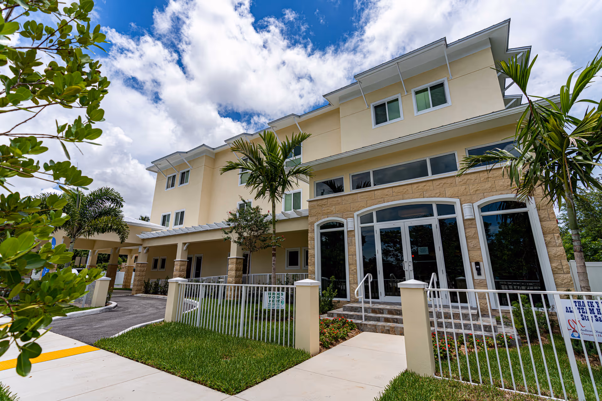 Front entrance of a three-story senior living building with stone accents, palm trees, and a gated walkway.