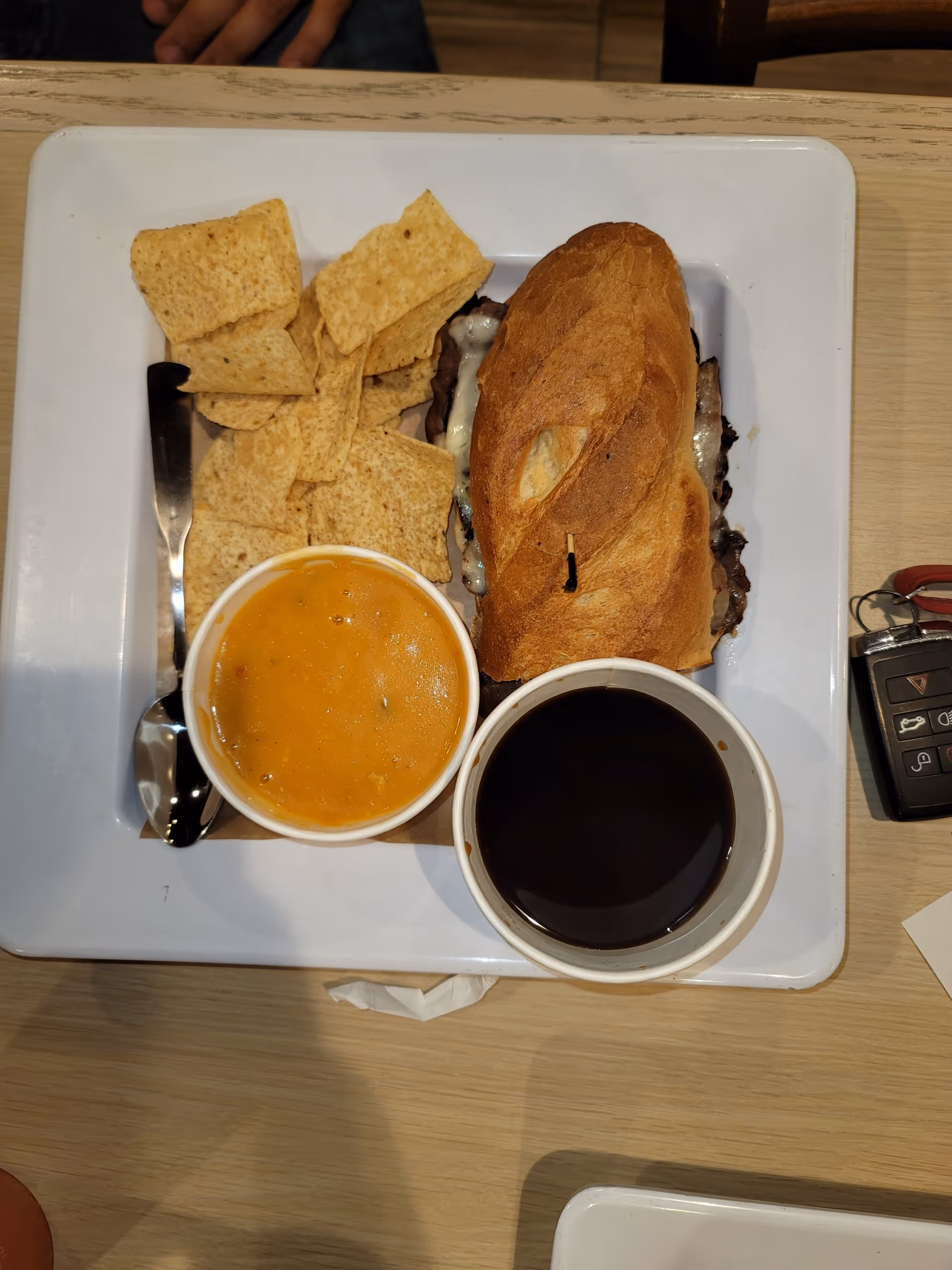 A tray on a table holding a sandwich, tortilla chips, and two bowls (one with a soup or stew and one with a dark liquid) with utensils and keys nearby.