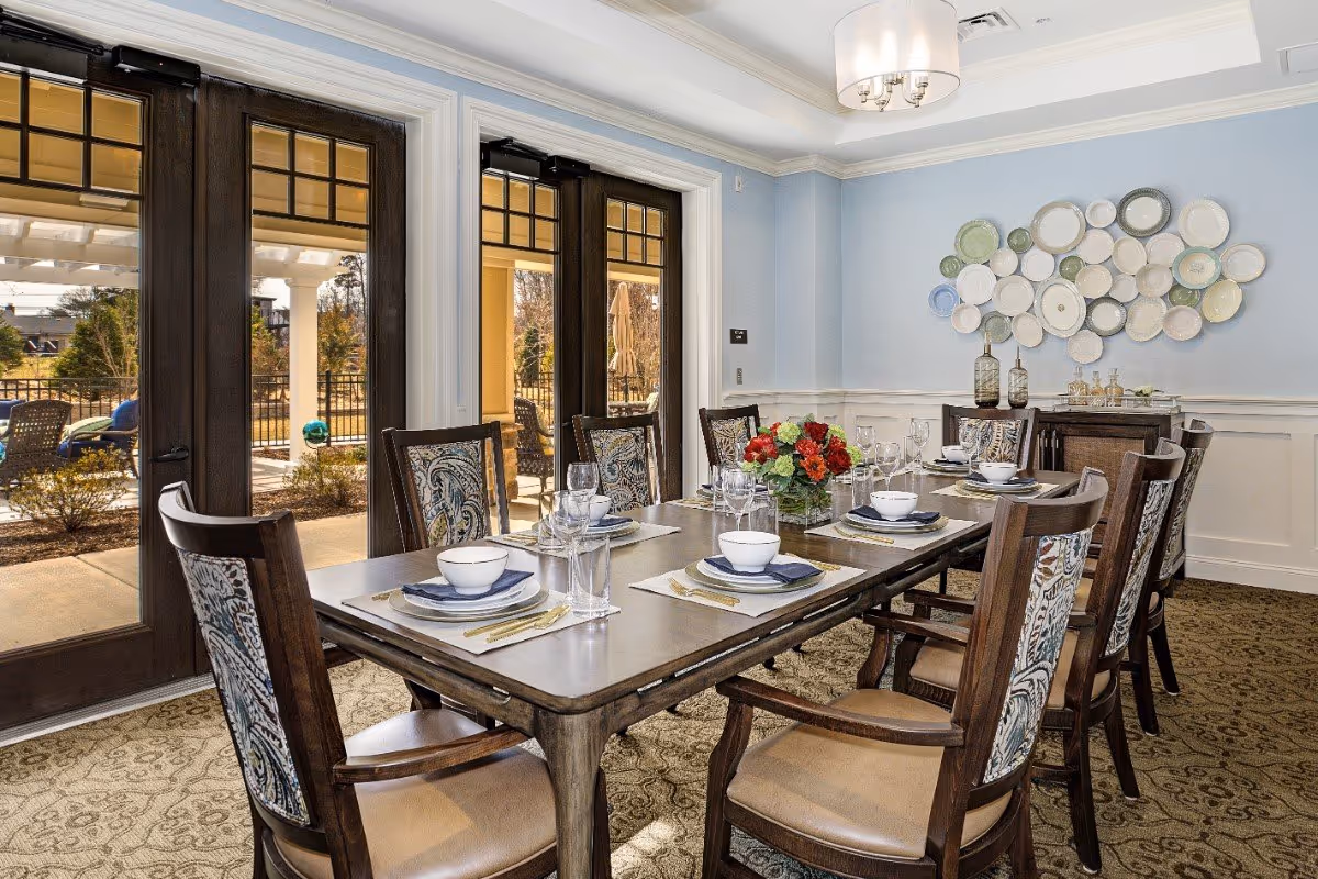 Dining room with a long wooden table set for a meal, upholstered chairs, decorative wall plates, and glass doors opening to an outdoor patio.
