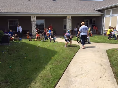 Children and adults gathering on a grassy courtyard outside a single-story assisted living building, searching for colorful eggs.